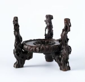  A small, dark brown stand with three legs designed to look like intertwined branches, featuring a flat, circular top with a decorative border and an intricately carved, hollow space below it.