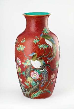  An intricately designed vase with a glossy, cherry-red background featuring hand-painted scenes of birds among branches and flowers, predominantly in whites, browns, blues, greens, pinks, and yellows, with turquoise edging at the opening and base.