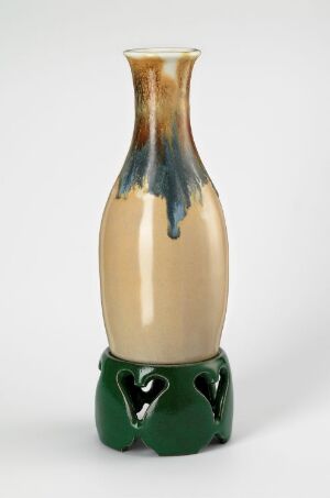  A two-part ceramic vase with a cream-colored upper section featuring a dark blue to teal drip glaze, and a dark green cylindrical stand with heart-shaped cutouts. Artist name and title unknown.