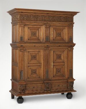  Antique two-part wooden cabinet on casters, with intricately carved panel doors in a medium-brown wood, detailed with metal hardware, emanating a sense of historical craftsmanship.