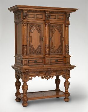  An elegant historical wooden cabinet with detailed geometric patterning on the doors, ornamental cornice, and turned legs on a raised base, set against a plain light grey background.