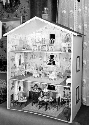 A black and white photograph of a detailed, three-story dollhouse with visible interior rooms, including an attic, bedroom, living area, and dining room, each meticulously furnished and decorated.