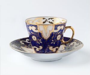  A hand-painted Feldspar porcelain cup and saucer set by Porsgrunds Porselænsfabrik AS, featuring an ornate design with a deep blue background, gold and white decorative elements, and a wavy edged saucer with gold trim.