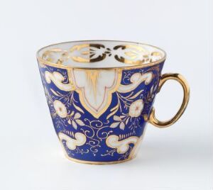  Porcelain cup by Porsgrunds Porselænsfabrik AS with a rich cobalt blue background and hand-painted overglaze decoration in enamel colors with symmetrical gold detailing, shield-like emblem, curling leaves, and flowers, complete with a gilded handle and rim.