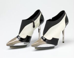  A pair of high-heeled shoes with a bold black and white panel design and shiny metallic silver toe caps and heels, featuring a center black zipper for closure, set against a light background.