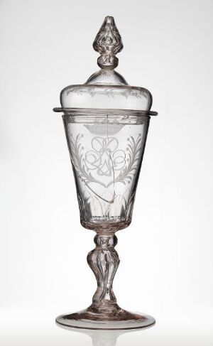  An intricately etched glass goblet with a matching lid, featuring delicate floral motifs, standing against a light background, showcasing its transparent elegance and fine craftsmanship.