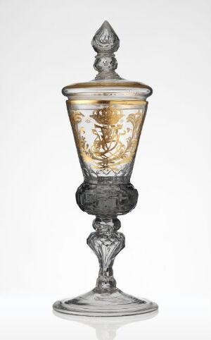  An ornate glass goblet with a gold accented bowl and a lid with a pointed finial, set against a white background. The stem and base of the goblet have a metallic silver appearance, adding to its decorative quality.