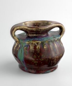 
 A small, ancient-looking ceramic pot with iridescent blue and green glazing on the upper rim and handles, set against a deep brown base color.