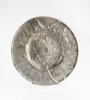  A silver-toned decorative plate with embossed floral patterns, displayed against a plain white background. Artist and title unknown.