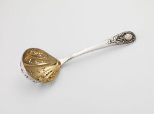  An ornate silver tea strainer spoon has a detailed handle and a pear-shaped bowl with a patterned strainer, sitting against a plain light background. Artistname and title are unknown.