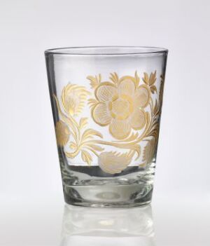  An empty transparent glass with a golden floral pattern etched onto its side, standing on a white background. The glass has a thick base and tapers slightly upwards, and the design features full-petaled flowers with swirling stems and leaves.