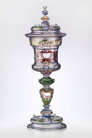  An elaborate covered glass vessel with a tiered silver-blue pedestal, emerald green and rose pink sections on the stem, a cylindrical body featuring a white paneled scene with floral accents, and a translucent lavender-tinted lid topped with a multicolored finial.