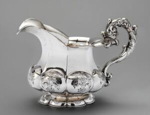  An ornate silver creamer with embossed floral motifs and a sculpted, fantastical creature for a handle, set against a light gray background.