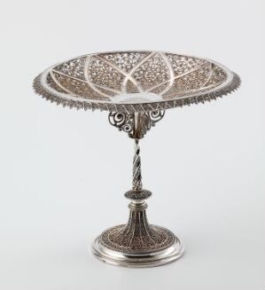  An intricate silver compote with a lacy, filigree patterned tray on top of a slender pedestal, displaying elaborate craftsmanship and sitting against a plain white background.