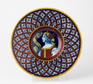  Decorative plate featuring a central portrait of a regal figure in profile, surrounded by a geometric pattern of white diamonds with red and blue dots on a blue background and a golden yellow border.