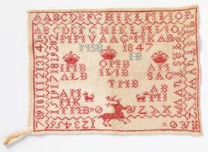  An off-white traditional sampler with red embroidery featuring two sets of alphabets, numbers, symmetrical floral patterns, and a scene with deer at the bottom, bordered by a simple stitched line with a tassel on one corner.