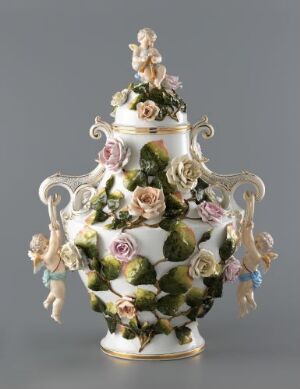  An ornate porcelain vase with a bulbous body, decorated with pastel-colored sculpted flowers, green leaves, and gold accents. It has graceful handles and a small figurine on the lid.