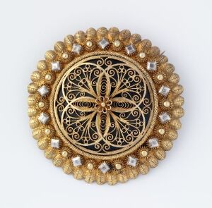  An ornate circular brooch with a textured gold beaded border, white diamond-shaped accents, and a central black and gold filigree pattern.