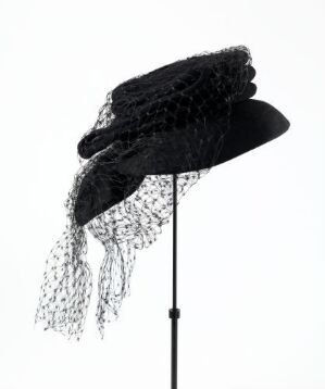  A black vintage-style hat with a downward-tilted brim and an elegant black netted veil, displayed on a simple hat stand against a white background. The image is monochromatic, emphasizing the classic design and texture of the hat.