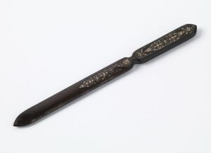  A dark brown, possibly wooden letter opener with light-colored, ornate decorations on its handle, lying on a light background. Artist name and title are unknown.