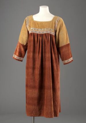  A traditional, rustic-style dress with a checkered pattern in rusty orange and reddish-brown, featuring an ornate cream-colored embroidered neckline, displayed on a featureless mannequin against a neutral background.