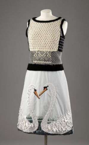  Julie Skarland's dress "Sail on" displayed on a mannequin, featuring a cream wool knitted top with dark geometric patterns, a middle section with sheer white fabric and embroidered swan figures, and a lower section with transparent overlay and sequin details against a neutral gray background.