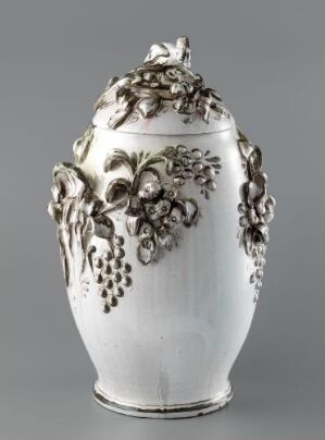  An ornate white urn with a lid, detailed with embossed grapevine motifs in a muted silver tone, against a light gray background.