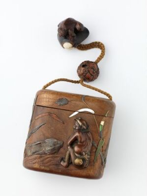  A traditional Japanese carved wooden box with intricate designs depicting a fisherman sitting and fishing beside reeds, with a large fish jumping from the water, all in a polished brown wood with variations in tones and textures, and a cord with two beads for carrying.