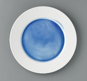  "Monokromer," a porcelain art piece by Heidi Bjørgan, featuring a circular dish with a vivid blue glazed center surrounded by a pristine white border, exhibiting the contrast and depth achieved through the turning and glazing technique.