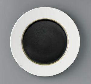  A circular porcelain dish titled 'Monokromer' by an unidentified artist, featuring a sleek design with a pure white outer rim and a glossy black center, accentuated by a thin, dark greenish-black line at the edge of the black glaze.