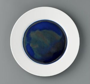  A porcelain art piece titled "Monokromer" by Anne Thomassen, featuring a deep blue and black glaze in the center, surrounded by a contrasting white rim. The design is simple and focuses on the interplay of dark, rich colors within a circular depression at the center of the round object, which has an overall effect of abstract and refined elegance.