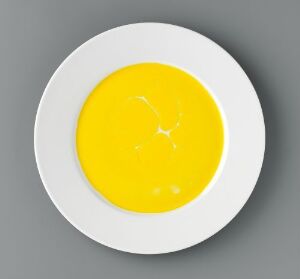  "Monokromer" by Heidi Bjørgan, a porcelain artwork resembling a fried egg with a glossy yellow glaze in the center on a white plate, set against a gray background.