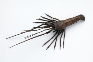  A sculptural object with a brown segmented body and numerous thin, dark metallic appendages radiating from the sides, displayed against a light gray background, evoking the likeness of an ancient sea creature or arthropod.