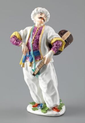  A porcelain figurine of a man with white hair and mustache, wearing traditional white clothing with a decorative multicolored vest and sashes, playing a small brown stringed instrument, standing on a base resembling grass with flowers.