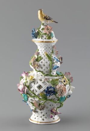  An elaborate porcelain sculpture featuring an array of colorful, detailed flowers, leaf patterns, and a bird, set against a light grey background. The sculpture exhibits a harmony of pastel pinks, soft purples, vibrant greens, and subtle yellows with a touch of blue dotted pattern over a white base.
