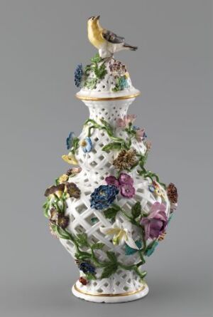  An intricately designed porcelain flask with a white lattice body, embellished with richly colored three-dimensional flowers and foliage, topped with a small porcelain bird figure, against a plain light grey background.