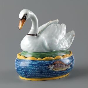  A porcelain figurine of a white swan with its wings slightly spread sitting on a vibrantly blue water-themed base with a painted fish and aquatic plants, all against a neutral grey background.