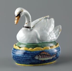  A ceramic container shaped like a swan on a water-like base, the swan is white with a black and orange beak, sitting on a blue and green base with a painted brown boat.