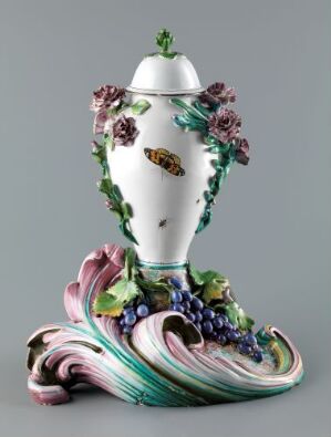  An ornate, baroque-style porcelain vase with white glossy finish, featuring pastel green and pink swirling base designs, adorned with three-dimensional purple and pink flowers, green leaves, dark purple grapes, and a golden emblem on its neck, culminating in a detailed lid with a handle.