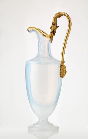  An elegant frosted glass pitcher with a golden, ornate handle against a soft white background. The pitcher's body has a delicate, translucent appearance, and the handle exhibits intricate details, indicating a luxurious and possibly antique piece.