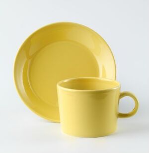  A set of yellow tableware with a shiny glaze, including a plate with a wide rim leaning in the background and a mug with a curved handle in the front, designed by Iittala.