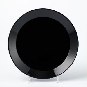  A sleek, black glazed flintware plate designed by Kaj Franck rests on a clear stand against a neutral background, showcasing its simple and elegant design with a reflective, glossy surface.