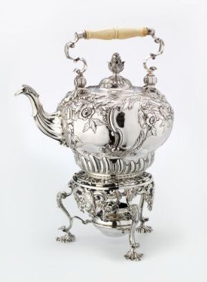  A silver kettle titled "Vannkjele" by Alexander Johnston, featuring ornate floral repoussé designs and a turned ivory handle, stands on a decorative silver stand with stylized legs, against a white background.