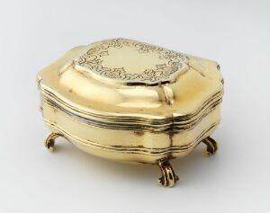  A small, golden-colored, ovular box with an etched design on the lid, standing on four elegant curved feet, set against a neutral grey background.