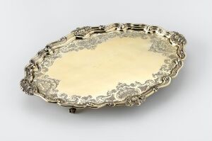  An oval-shaped silver serving tray with elaborate rococo scrollwork along its raised edges, featuring a reflective central surface and a warm, pale golden sheen.