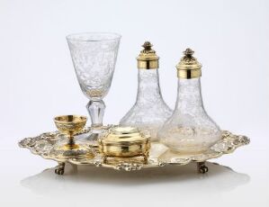  An elegant tableware set, including etched glass bottles with golden caps, a matching drinking glass, a small bowl, and a lidded container, all displayed on an ornately embossed metallic tray against a neutral background. Artist name and title: unknown.