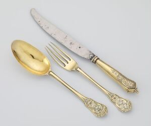  A set of golden and silver-colored cutlery, including an ornate spoon, fork, and knife with decorative handles, arranged on a neutral grey background.