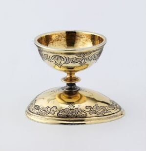  An ornate gold-tone chalice with intricate engravings on the exterior of the bowl and base, featuring a polished interior and a contrasting dark stem knob, set against a light background.