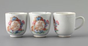  A trio of ornately painted porcelain mugs with a white base color, arranged in a row on a light-grey background. Each mug is decorated with a circular monogram and floral motifs in soft blues, pinks, oranges, greens, and touched with gold accents. The first mug faces forward, the second is turned slightly to show the side, and the third displays its side and a handle.