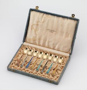 
 An open case displaying a set of vintage silver coffee spoons, "Mokkaskje," with gilded bowls and handles decorated with colorful enamel by an unidentified artist, against a cream-lined interior.